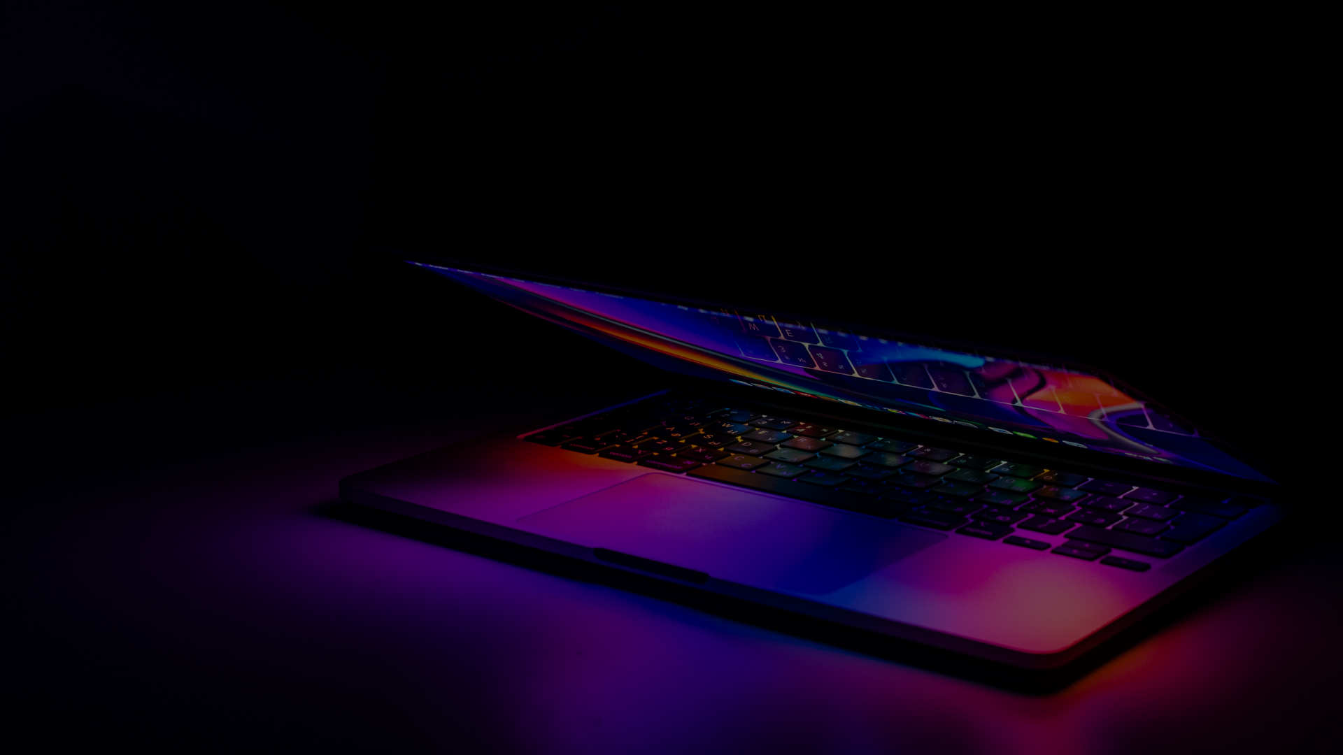 Un ordinateur portable entrouvert dans une pièce sombre, l’écran projetant une lumière colorée sur le clavier.